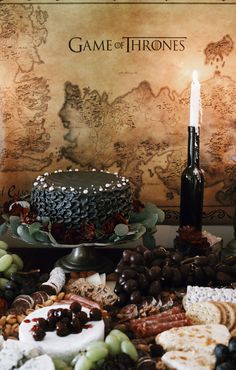 a game of throne cake surrounded by grapes, crackers and cheeses with a lit candle