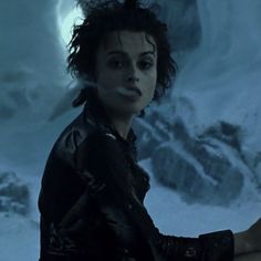 a woman with dark hair and black gloves standing in front of snow covered mountain side