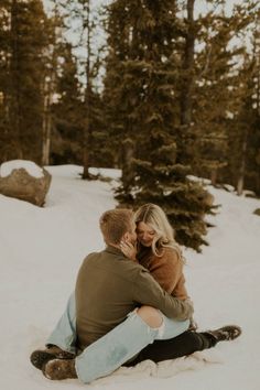 a man and woman sitting in the snow hugging each other with pine trees behind them