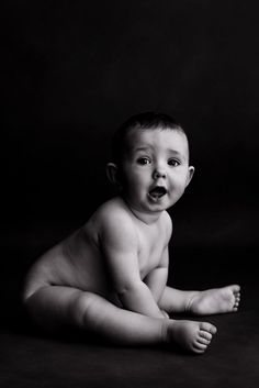 a black and white photo of a baby sitting on the floor with his tongue out