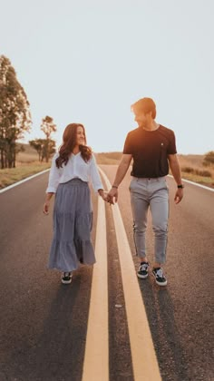 a man and woman holding hands walking down the middle of an empty road with trees in the background