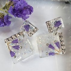 two square shaped purple and white crystal earrings on a table next to some purple flowers