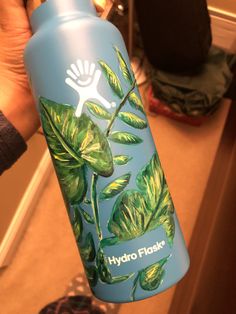 a hand holding a blue hydro flask with green leaves painted on the front and side