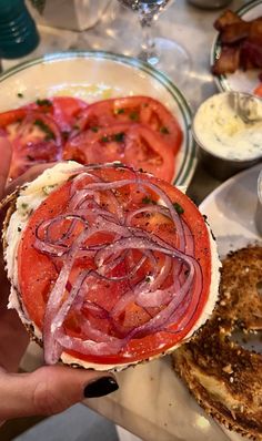 a person is holding up a sandwich with onions and tomato on it while sitting at a table full of food