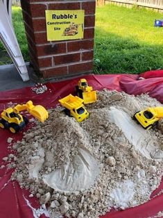 a red table topped with lots of sand next to a brick wall and toy trucks