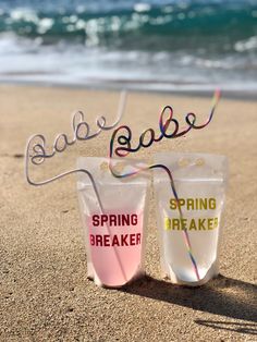 two plastic cups sitting on top of a sandy beach next to the ocean with writing