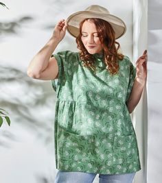 a woman wearing a hat and green top standing next to a white wall with her hands on her head