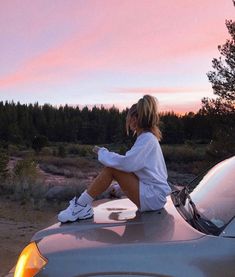 a woman sitting on the hood of a car in front of a forest at sunset