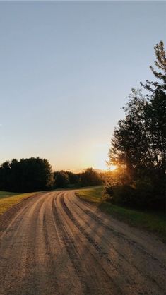 a dirt road with trees on both sides and the sun setting in the distance behind it