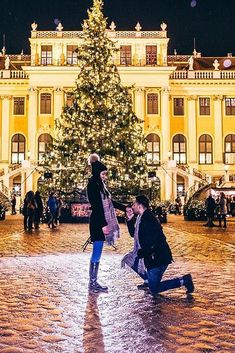 two people kneeling down in front of a christmas tree with lights on the building behind them