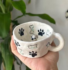 a hand holding a white dog paw print coffee cup in front of a potted plant