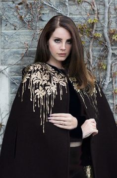 a woman wearing a black cape with gold beading on her shoulders and arms, standing in front of a brick wall