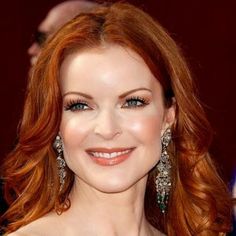 a woman with red hair and blue eyes smiles at the camera while wearing large earrings