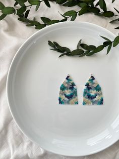 two beaded earrings sitting on top of a white plate next to green leafy branches