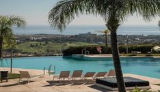 an empty swimming pool with lounge chairs and palm trees in the foreground, overlooking the ocean