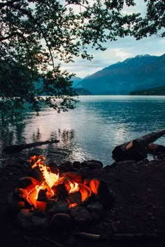 a campfire is lit on the shore of a lake with mountains in the background