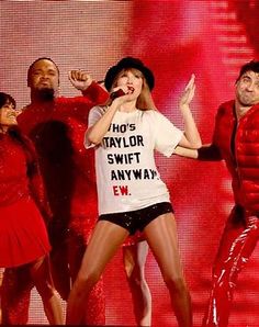 taylor swift performs on stage with dancers in red outfits and black hats, while holding her hands up