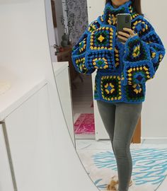 a woman standing in front of a mirror holding a cell phone and wearing a crocheted jacket