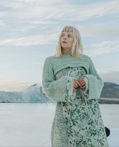 a woman standing in the snow wearing a green sweater and floral print dress with her arms folded