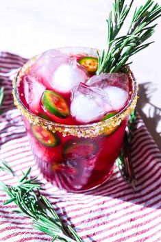 a red drink with ice and rosemary garnish in a glass on a striped cloth