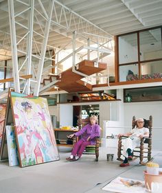 two people sitting in chairs with paintings on the floor and stairs to the second floor