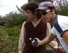 two young men standing next to each other in front of purple flowers and bushes, one holding a cell phone