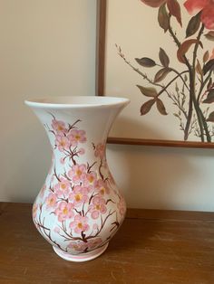 a white vase sitting on top of a wooden table next to a framed flower painting