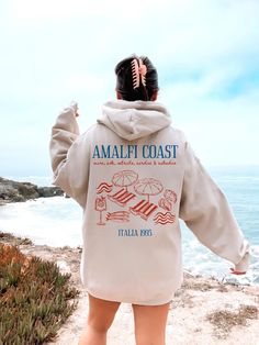 a woman standing on the beach with her back to the camera, wearing an amalfi coast sweatshirt