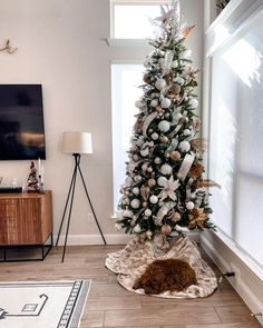 a christmas tree with white and gold ornaments in a living room area next to a window