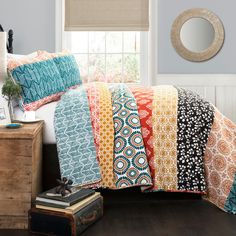 a bed covered in lots of different colored sheets and pillows next to a window with a mirror