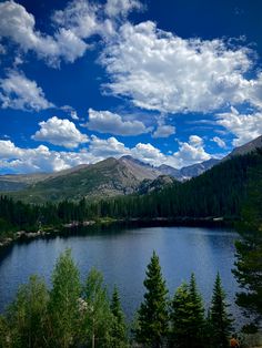 a lake surrounded by trees and mountains under a blue sky with white puffy clouds