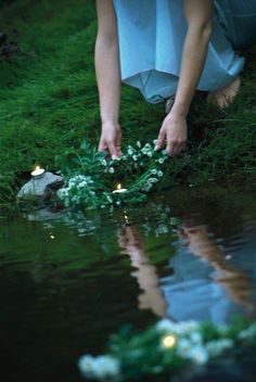 a woman kneeling down next to a body of water with flowers in her hands on the ground