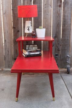 a red table with books and a lamp on it in front of a wooden fence