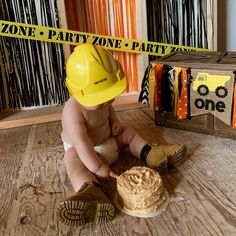 a baby sitting on the floor wearing a hard hat and holding a cake in front of him