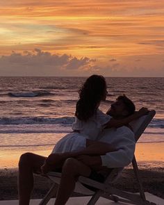 a man and woman sitting on top of a beach chair next to the ocean at sunset