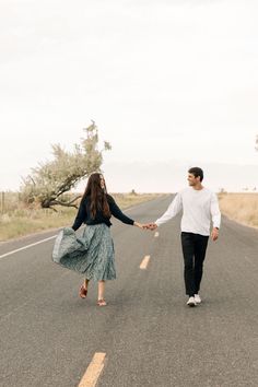 a man and woman holding hands walking down the middle of an empty road with trees in the background