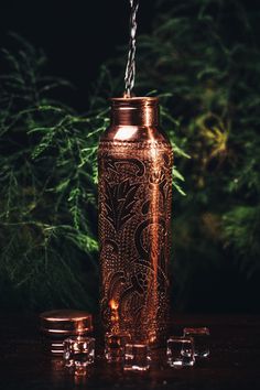 a copper colored mason jar with two glasses next to it and some green plants in the background