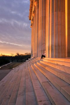 a person sitting on steps in front of the lincoln memorial at sunset, with columns lit up