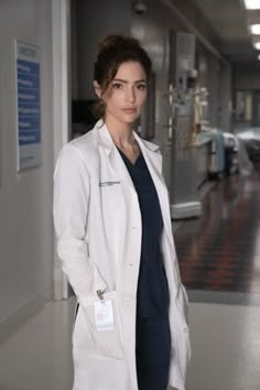 a woman standing in a hospital hallway wearing a white coat and blue scrubs with her hands on her hips