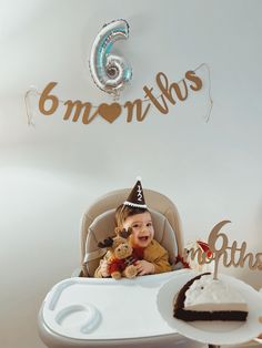 a baby sitting in a high chair with a birthday cake and balloons on the wall