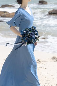 a woman in a long blue dress holding a bouquet on the beach near the ocean