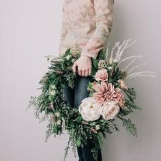 a woman holding a wreath with flowers and greenery around her waist, against a white wall