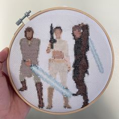 a hand holding a cross stitched picture of three people in star wars outfits with swords