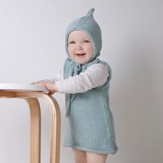 a small child wearing a blue knitted sweater and hat standing next to a wooden chair