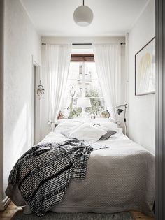 a bed sitting under a window next to a white wall and wooden floor covered in blankets