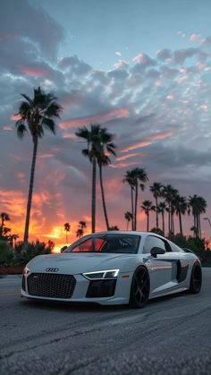 a white sports car driving down a street next to palm trees at sunset with the sun setting in the background