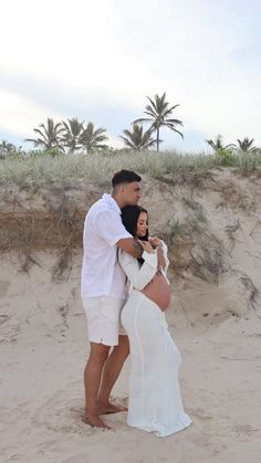 a man and woman standing on top of a sandy beach next to the ocean with palm trees in the background