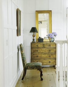 a chair sitting in front of a dresser next to a mirror and lamp on top of a wooden floor