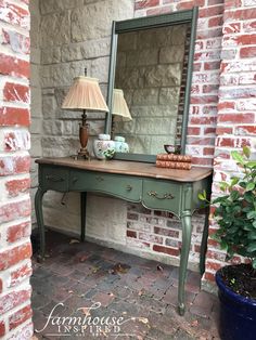 a green table with a mirror and lamp on it next to a brick wall in front of a potted plant