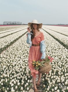 a woman standing in a field with flowers wearing a hat and holding a wicker basket